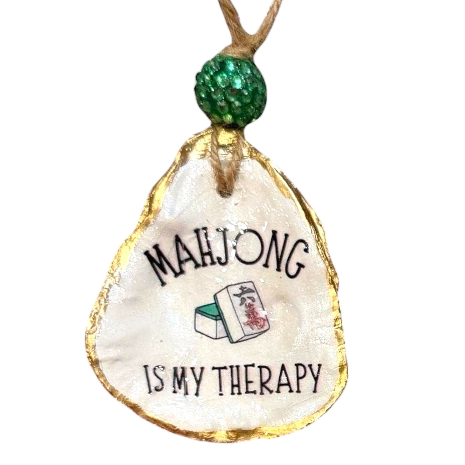 A decorative hanging ornament shaped like an oyster shell, with a gold-leaf textured border, a green glitter bead at the top, and the printed phrase 'MAHJONG IS MY THERAPY' with a small mahjong tile illustration.