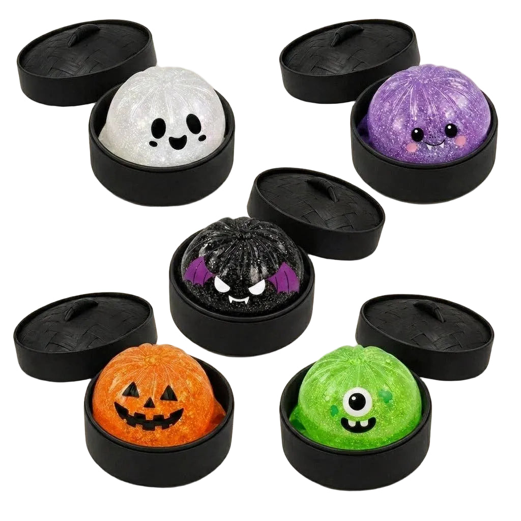 Five Halloween-themed squishy bath bombs in round black containers with lids, arranged on a beige background with bat and spiderweb illustrations. The bath bombs are white with black eyes, purple glitter, black glitter with purple ears, orange jack-o'-lantern, and green with a single eye.