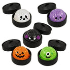 Five Halloween-themed squishy bath bombs in round black containers with lids, arranged on a beige background with bat and spiderweb illustrations. The bath bombs are white with black eyes, purple glitter, black glitter with purple ears, orange jack-o'-lantern, and green with a single eye.