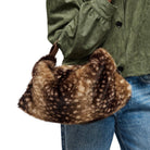 A person wearing an olive green jacket and blue jeans holds a brown and beige spotted faux fur shoulder bag against a white background.