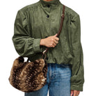 A person wearing an olive green long-sleeve button-front jacket and blue jeans, holding a brown faux-fur shoulder bag with a brown strap against a plain white background.
