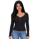 A woman modeling a black long-sleeve henley top with silver buttons down the front, paired with blue jeans, against a plain white background.