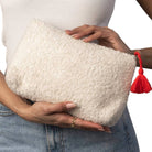 A person holds a cream-colored, textured fleece clutch bag with a red tassel zipper pull. The person is wearing a white ribbed top and blue denim jeans, with a gold ring on one finger, against a plain white background.