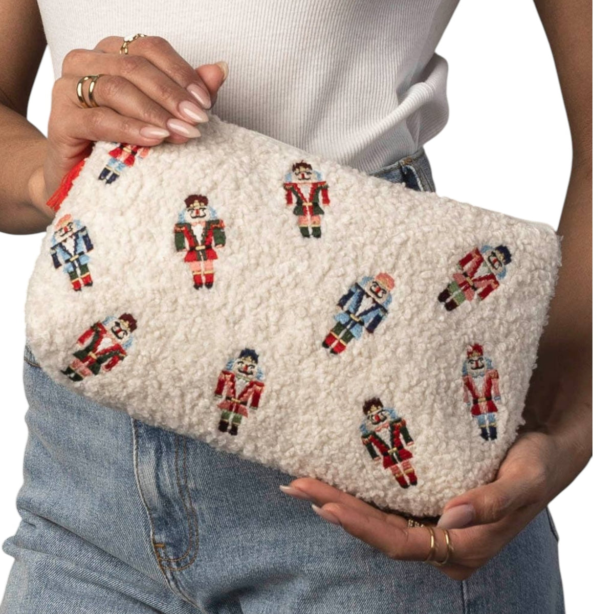 A person holds a cream-colored, sherpa-textured clutch bag decorated with a repeating pattern of embroidered nutcracker soldiers in red, blue, and green. The person is wearing a white ribbed top and light-wash blue jeans.