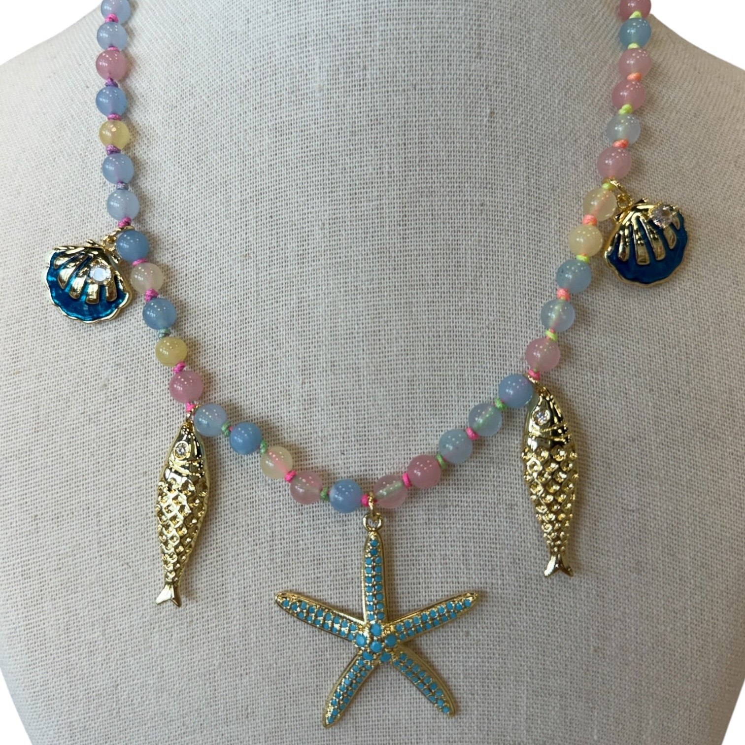 A beaded necklace displayed on a fabric mannequin bust, featuring pastel pink, blue, and lavender beads with gold-tone sea-themed charms including a starfish, two fish figures, and two seashells.