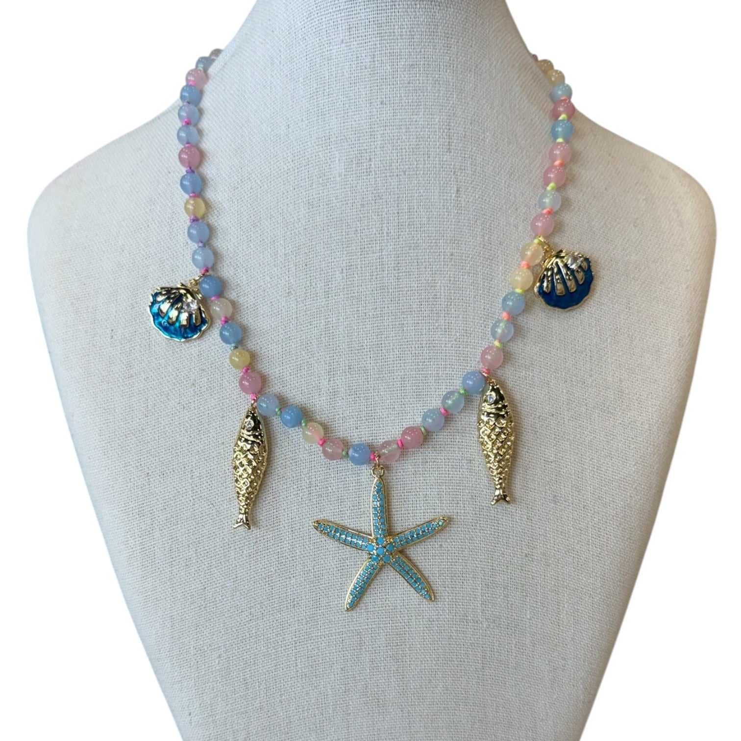 A pastel beaded necklace with seashell and starfish charms displayed on a cream fabric mannequin bust. The necklace features light blue, pink, and yellow beads, gold-toned fish charms, and a turquoise starfish pendant.