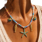 A close-up of a person wearing a colorful beaded necklace with gold-toned shell and fish charms, plus a turquoise starfish pendant, against a white lace top.