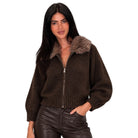 A woman modeling a dark brown knit zip-up jacket with a fuzzy brown faux-fur collar, paired with dark brown faux-leather pants, against a solid white background.
