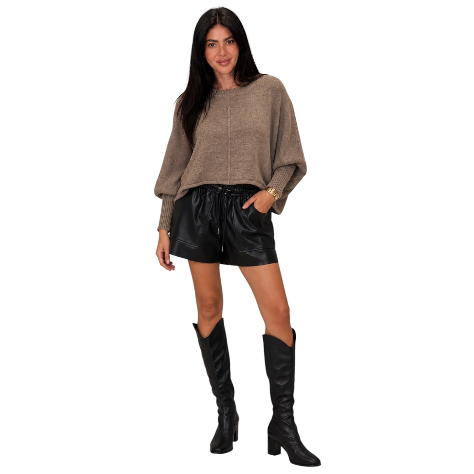 A woman modeling a taupe knit long-sleeve crop top, black faux-leather shorts, and black knee-high boots against a plain white background.