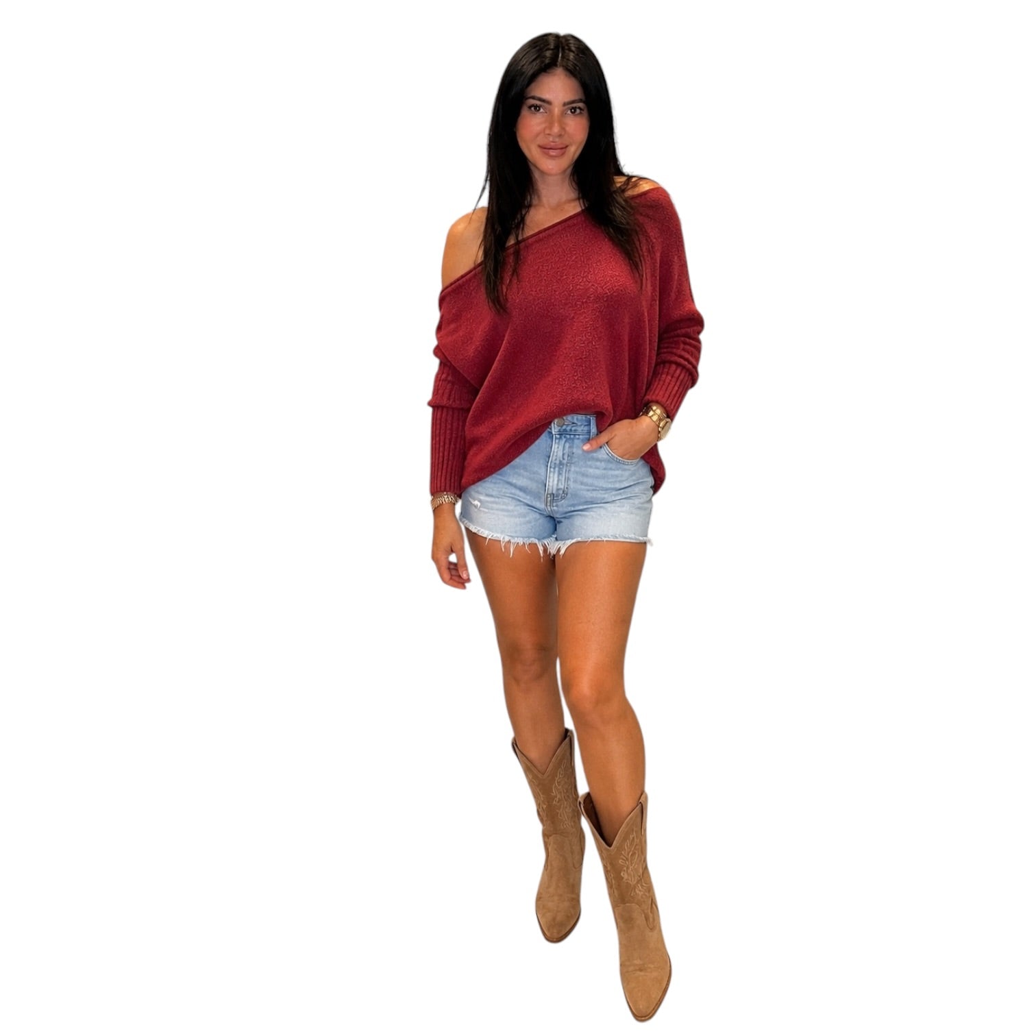A woman modeling a long-sleeve off-the-shoulder knit top in a deep burgundy red, paired with light-wash denim shorts and tan cowboy boots, set against a solid white background.