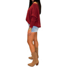 A woman shown from the side wearing a loose off-the-shoulder knit sweater in burgundy, light denim shorts, and tan ankle boots, against a plain white background.