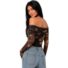 A woman wearing a sheer off-the-shoulder long-sleeve bodysuit with a black and brown floral lace pattern, paired with light-wash blue jeans, shown from a three-quarters back view against a solid white background.