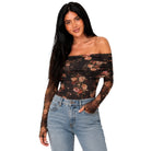 A woman modeling an off-the-shoulder long-sleeve bodysuit with a sheer black mesh and brown floral pattern, paired with light blue jeans, against a plain white background.