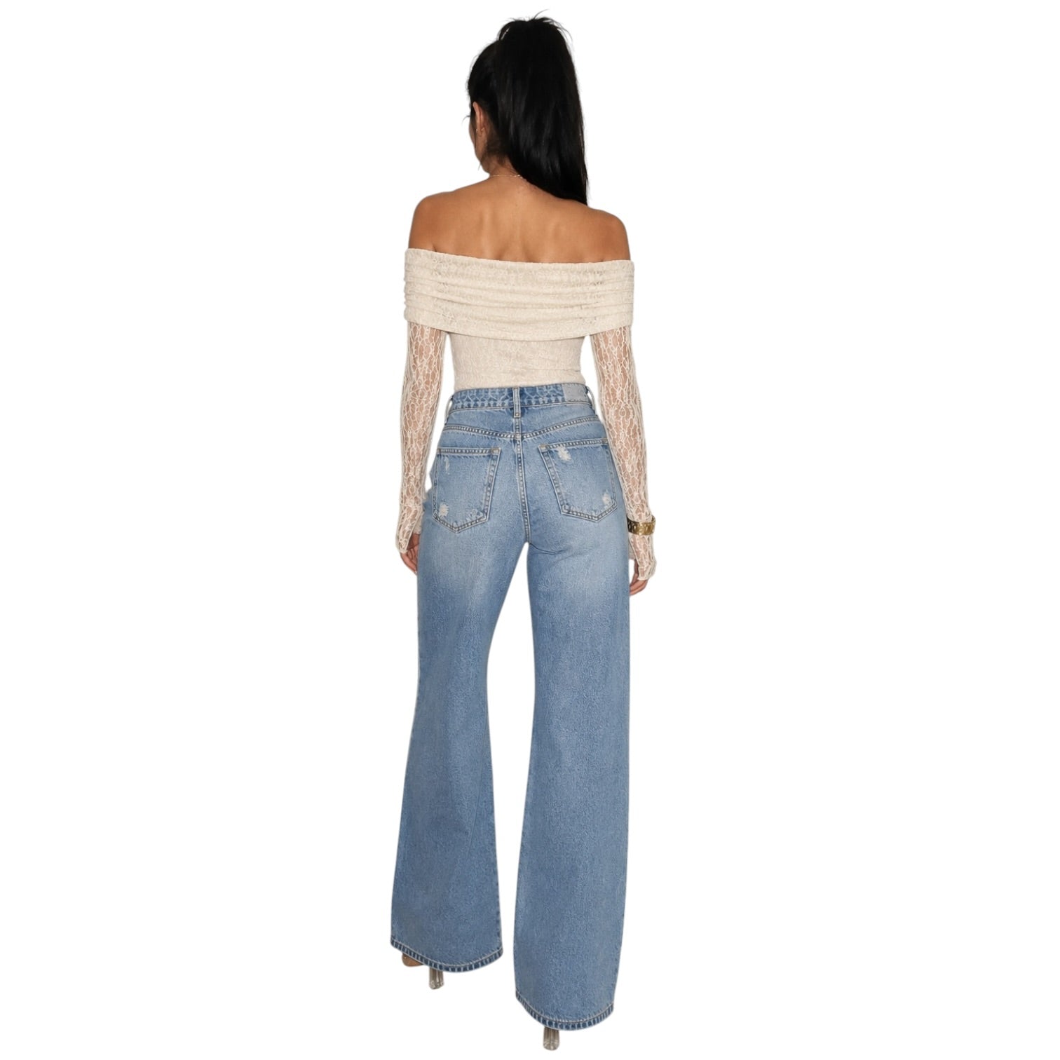 A woman shown from the back wearing a cream off-the-shoulder knit top with sheer long sleeves and light-wash blue wide-leg jeans, against a plain white background.