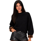 A woman modeling a black ribbed knit sweater with leopard-print faux fur cuffs, paired with black leather-look shorts, against a solid white background.