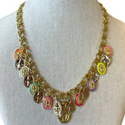 A gold-tone chain necklace displayed on a cream fabric mannequin bust, featuring multiple oval and round religious medallions in assorted colors including pink, orange, green, purple, yellow, and brown.