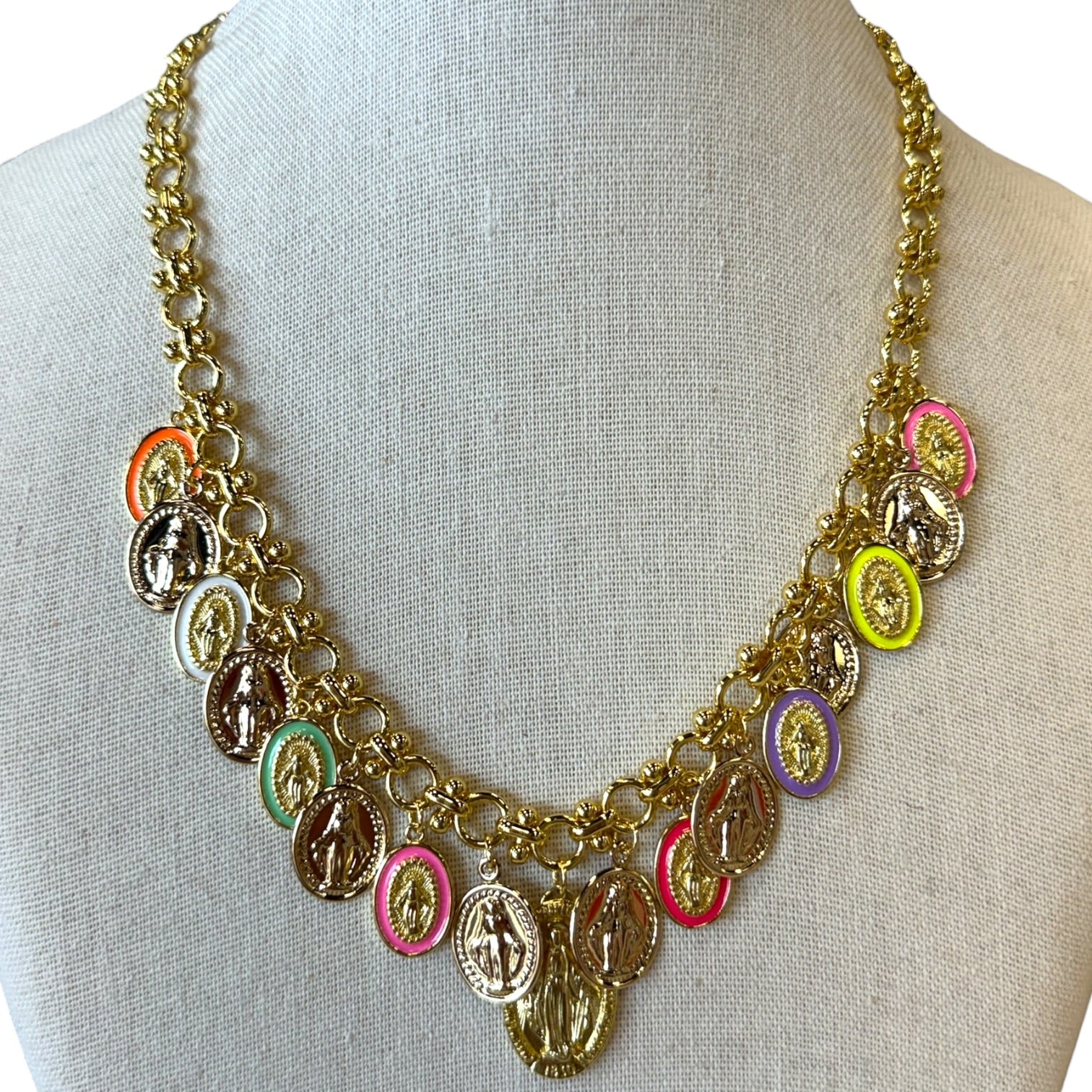 A gold-tone chain necklace displayed on a cream fabric mannequin bust, featuring multiple oval and round religious medallions in assorted colors including pink, orange, green, purple, yellow, and brown.