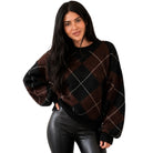 A woman modeling a brown and black argyle knit sweater with thin white diagonal lines, paired with black faux-leather pants, against a plain white background.
