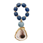 A beaded necklace featuring dark blue textured glass beads, a light blue stone bead, and a large natural oyster shell pendant with gold-toned metal trim, shown on a plain white background.