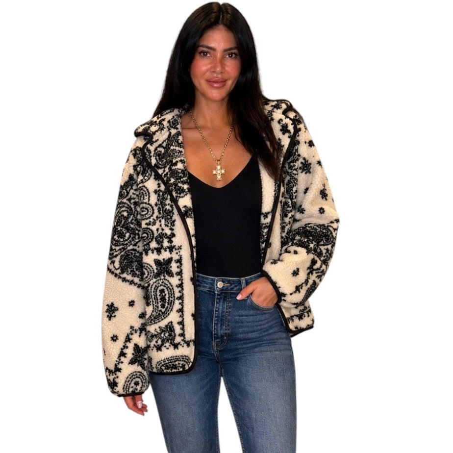 A woman modeling a black and cream paisley patterned zip-up jacket over a black top and blue jeans, photographed against a plain white background.