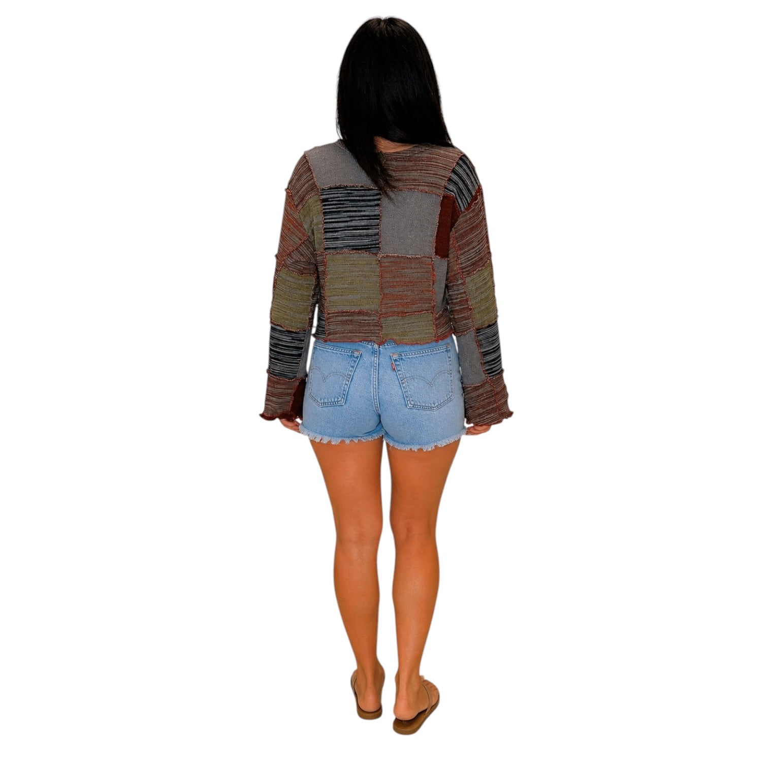 A woman viewed from behind wearing a long-sleeve patchwork knit top with horizontal striped panels in earthy tones, paired with light blue denim shorts and tan flat sandals, against a plain white background.