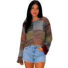 A woman modeling a long-sleeve patchwork knit top with horizontal striped panels in gray, brown, rust, olive, and beige, paired with light blue denim shorts against a plain white background.