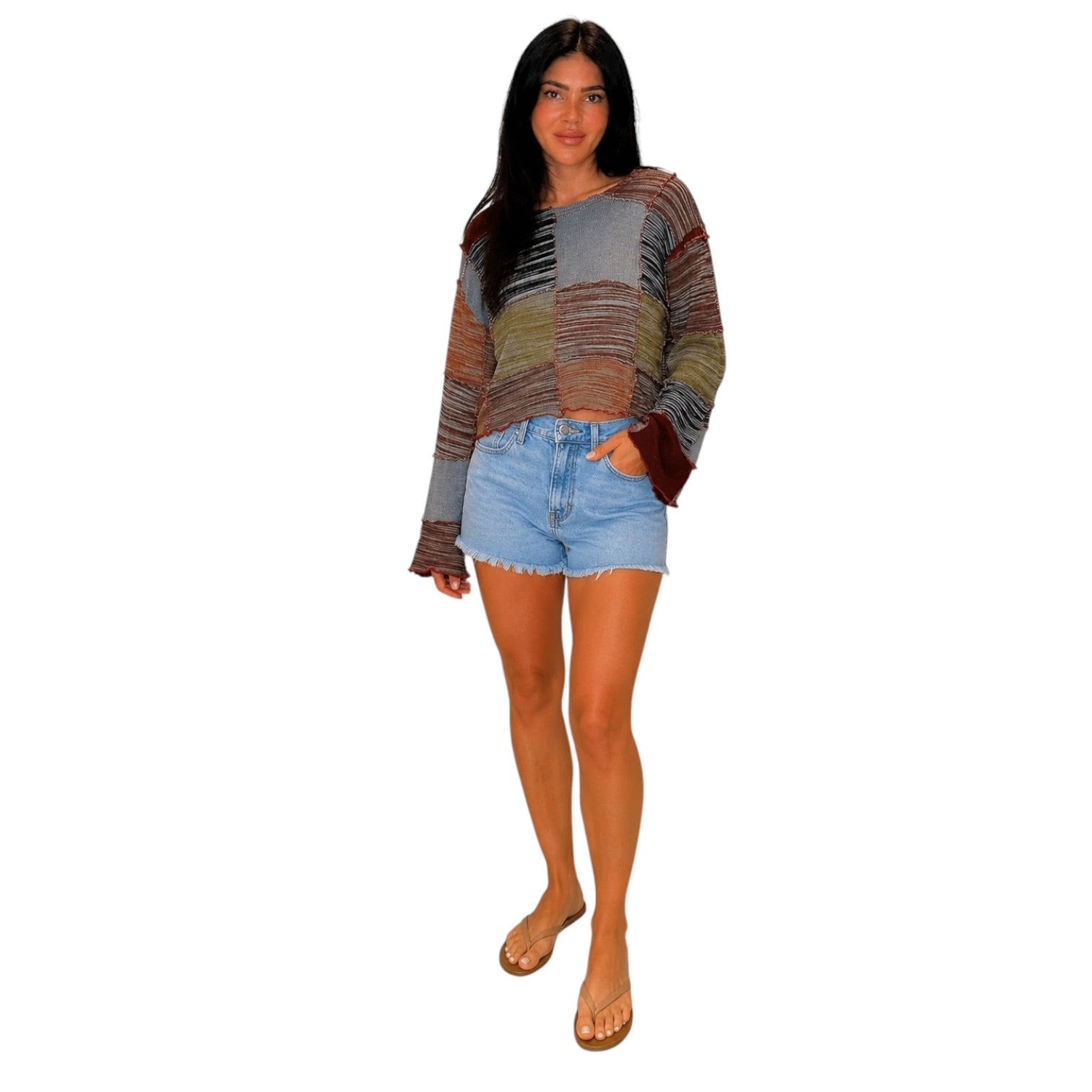A woman modeling a long-sleeve knit top with a multicolor patchwork pattern in shades of brown, gray, beige, and green, paired with light blue denim shorts and tan sandals, set against a plain white background.