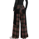 A person shown from the back wearing wide-leg patchwork pants with a plaid pattern in black, burgundy, and gold, featuring solid black patch pockets and a black corduroy back panel, paired with a black long-sleeve top and white sneakers.