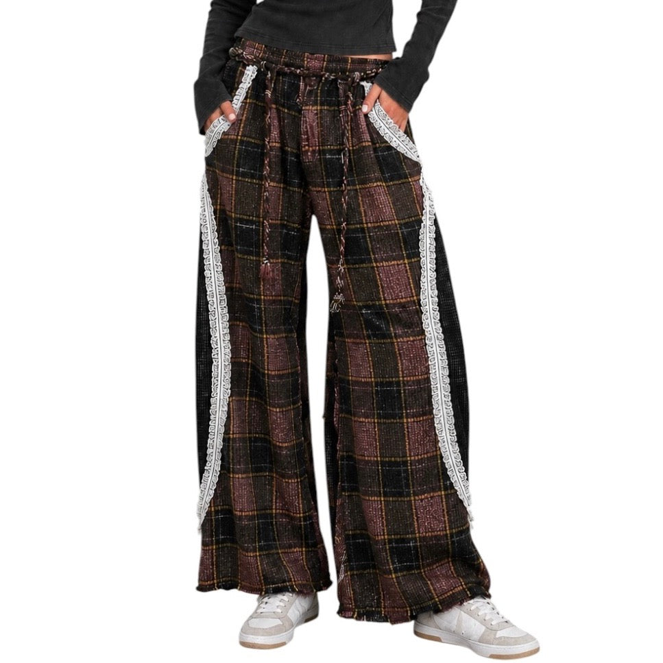 A person wearing loose, wide-leg plaid pants with black side panels and silver fringe trim, paired with a black long-sleeve top and white sneakers, shown against a plain white background.