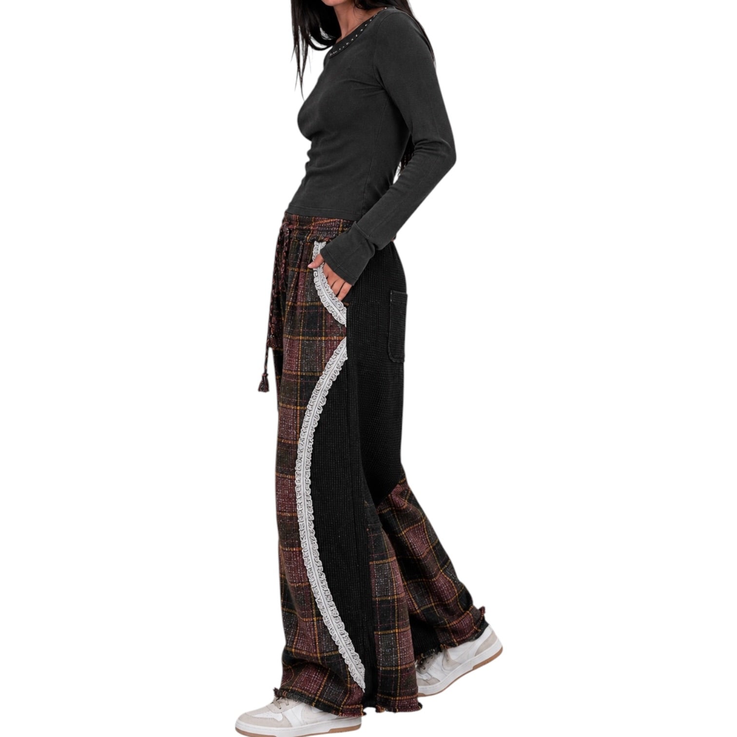 A model wearing a black long-sleeve top and wide-leg pants featuring a black base with a red and brown plaid side panel and a white lace-like vertical stripe.