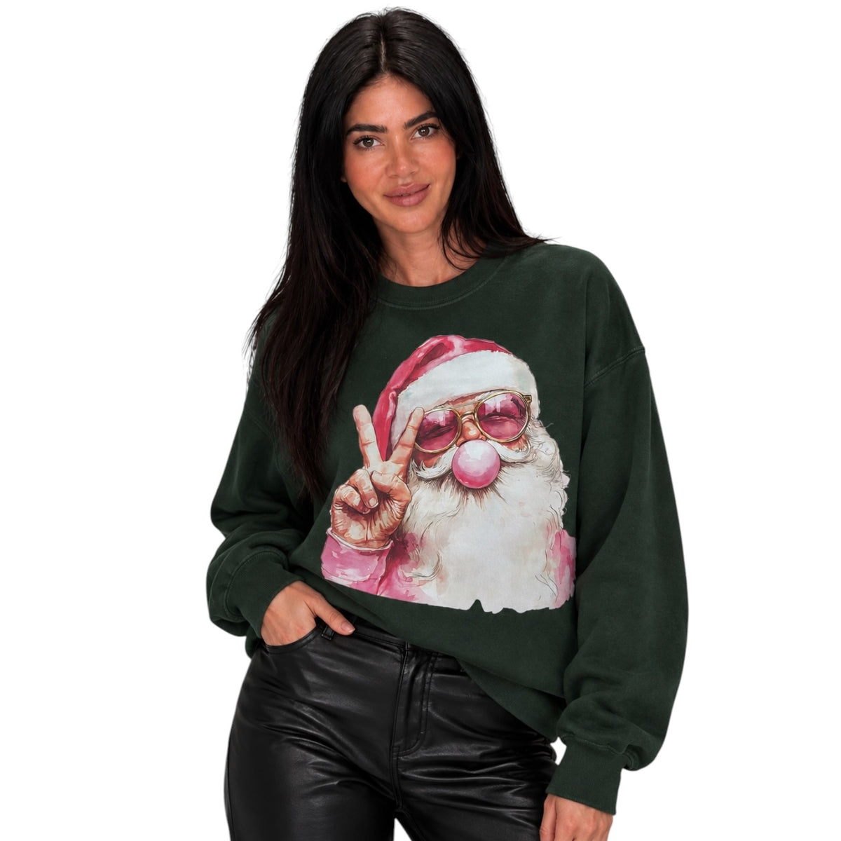A woman modeling a dark forest green crewneck sweatshirt featuring a graphic print of a Santa Claus character wearing pink sunglasses and making a peace sign. She is also wearing black leather pants against a solid white background.
