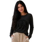 A woman modeling a black long-sleeve ribbed knit top with a large black peace sign graphic on the front, accented with small gold studs, paired with beige pants, against a solid white background.