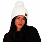 A woman wearing a white ribbed knit beanie with a fluffy pom-pom on top and a small brown brand patch on the side, photographed against a plain white background.