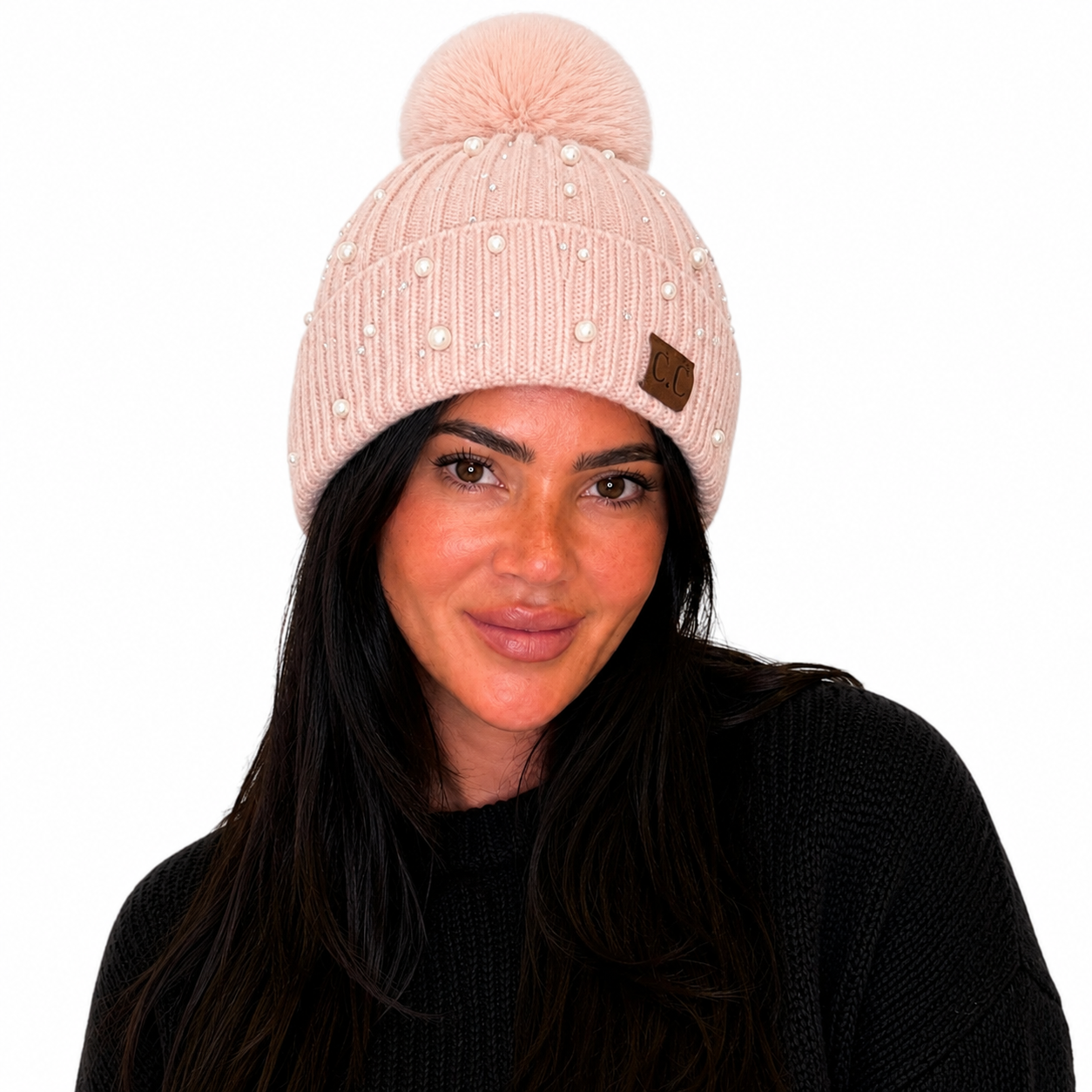 A woman wearing a light pink ribbed knit beanie with a matching pom-pom, decorated with small pearl-like embellishments and a small brown leather patch.