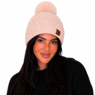 A woman wearing a light pink ribbed knit beanie with a matching pom-pom, decorated with small pearl-like embellishments and a small brown leather patch.
