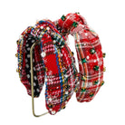 A pair of knotted fabric headbands with a festive Christmas theme. One headband features a red and black plaid pattern with white snowflakes, blue and yellow stripes, and white pearl beads, while the other features a red and white plaid pattern with green holly accents and small gold beads.