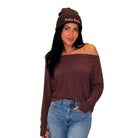 A woman modeling a dark brown off-the-shoulder long-sleeve top and a matching dark brown knit beanie with 'Petty Betty' embroidered in pink on the front, paired with blue jeans against a plain white background.