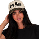 A woman wearing a cream-colored snapback cap with a leopard-print brim and 'Petty Betty' embroidered in black script on the front, paired with a black t-shirt against a solid white background.