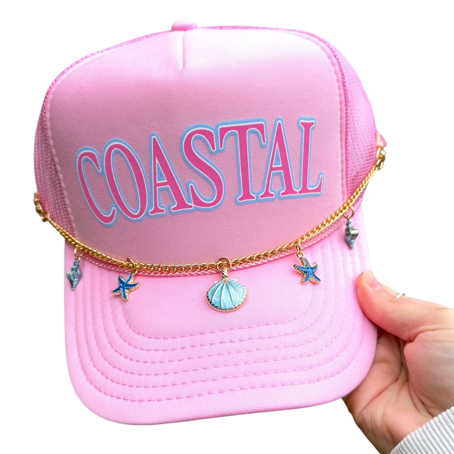 Pink cap with 'COASTAL' text and charms held by a hand on a white background