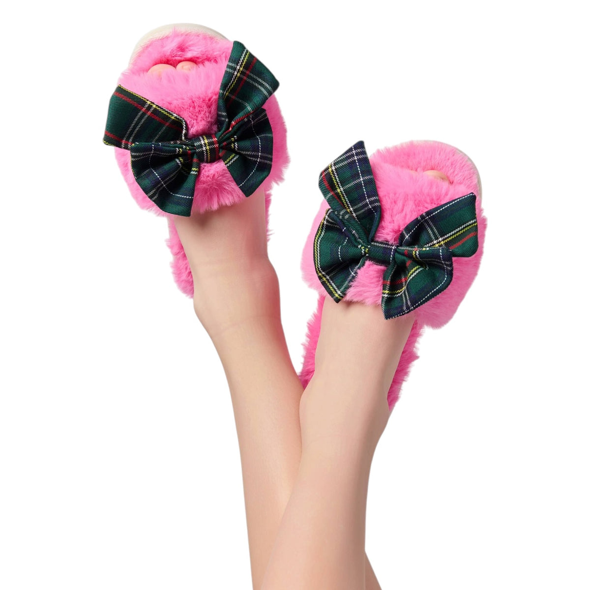 A pair of fluffy pink slip-on slippers with large green plaid bow details, shown being worn on feet against a plain white background.