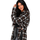 A woman modeling a brown, black, and white plaid button-up shirt with a fringed hem, worn open over a black crop top and black faux-leather pants, against a solid white background.