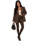 A woman modeling a brown plaid blazer, brown leggings, a white top, and dark brown ankle boots, holding a dark brown textured handbag against a plain white background.