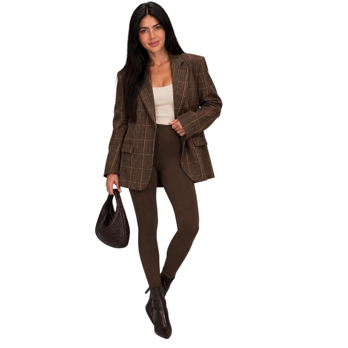 A woman modeling a brown plaid blazer, brown leggings, a white top, and dark brown ankle boots, holding a dark brown textured handbag against a plain white background.
