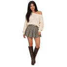 A woman modeling an off-the-shoulder cream knit sweater, a gray pleated mini skirt, and brown knee-high leather boots against a plain white background.