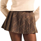 A pleated mini skirt in a brown and beige plaid pattern, worn by a model with a beige knit sweater against a plain white background.