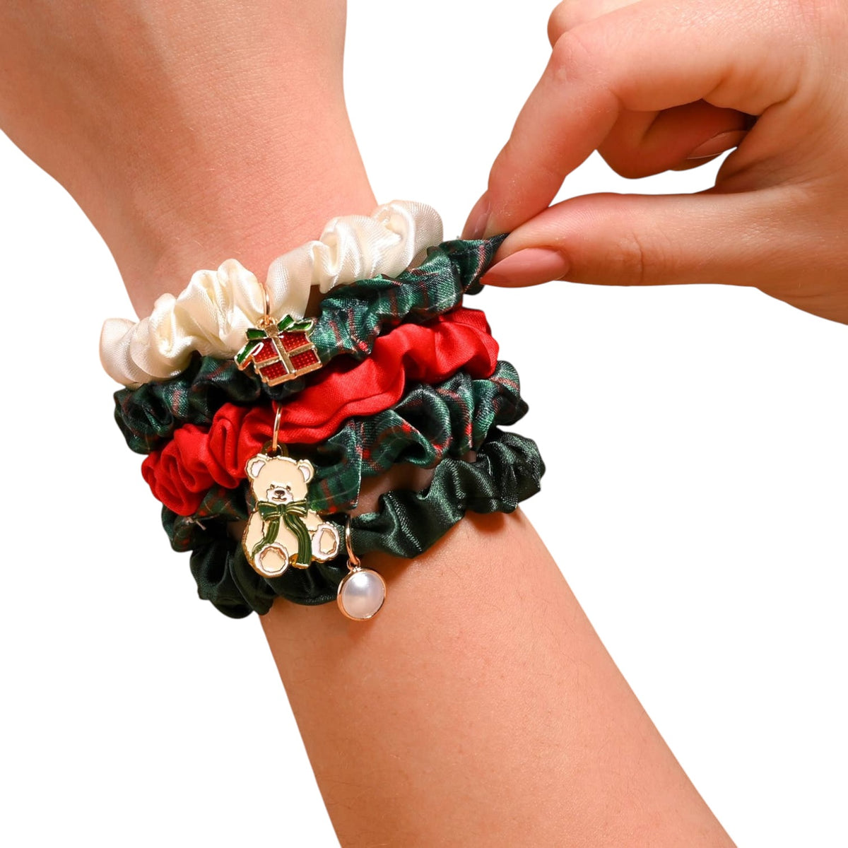 A close-up of a person's wrist wearing a stack of three satin scrunchies in white, red, and dark green plaid, each decorated with a small Christmas-themed charm (a gift box, a teddy bear, and a pearl). A hand is adjusting the top scrunchie against a plain white background.