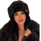 A woman wearing a dark brown faux fur headband with a small brown leather logo patch and a matching dark brown faux fur coat, set against a solid white background.