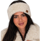 A woman wearing a fluffy cream faux-fur headband with a small brown leather logo patch that reads 'JC', paired with a matching cream faux-fur coat against a plain white background.