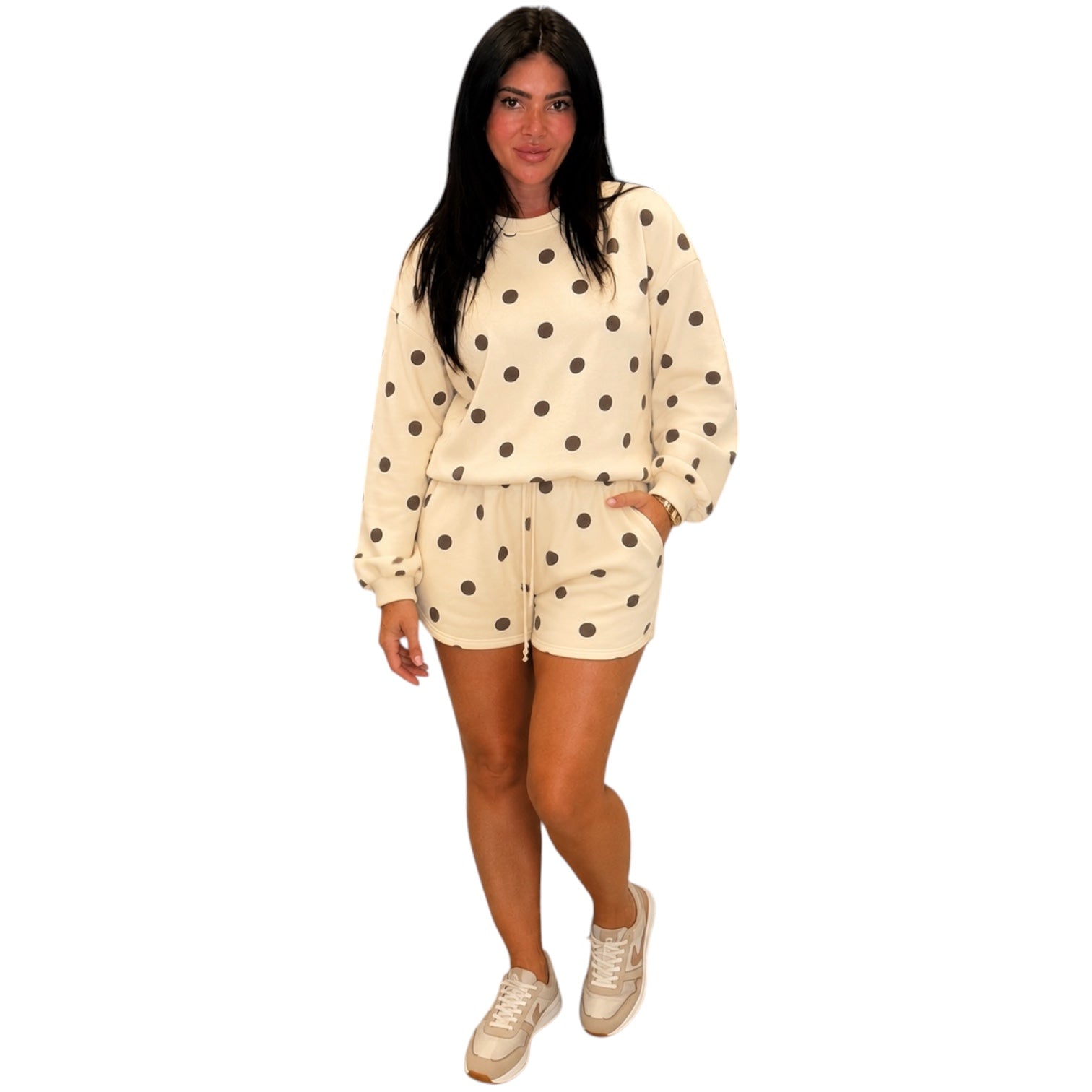 A woman modeling a matching cream-colored two-piece lounge set with dark brown polka dots, consisting of a long-sleeve sweatshirt and shorts, paired with beige sneakers on a solid white background.