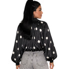 A woman shown from the back wearing a black satin-like blouse with large white polka dots and voluminous long sleeves, paired with light gray jeans, against a plain white background.